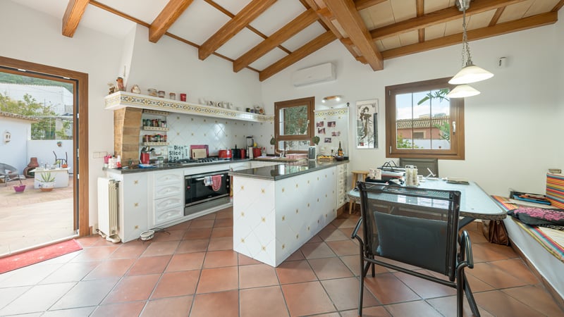 Mediterranean kitchen featuring terracotta floors, wooden ceiling beams, a white island, and a dining area by the terrace.