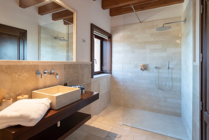 Modernt badrum med naturstensväggar, walk-in takdusch och synliga takbjälkar i trä.