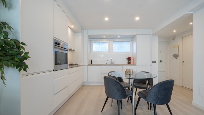 Clean white minimalist kitchen featuring handleless cabinetry, glass dining table, and grey velvet chairs.