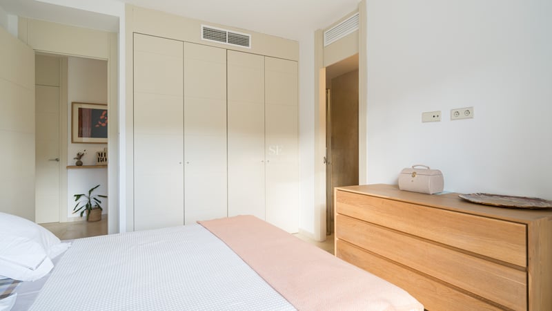 Ljust sovrum med krämfärgade garderober, träbyrå och säng med pastellrosa pläd.