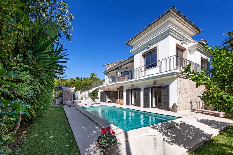 A white two-story villa with a turquoise swimming pool, sun loungers, stone walls, and lush greenery under a clear sky.