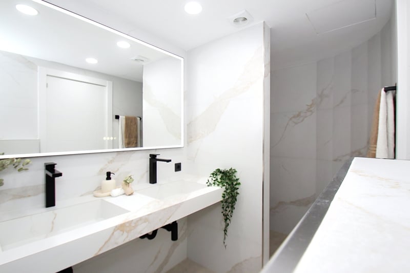 Ljust badrum med dubbelt tvättställ i vit marmor, mattsvarta kranar och en stor bakgrundsbelyst spegel.