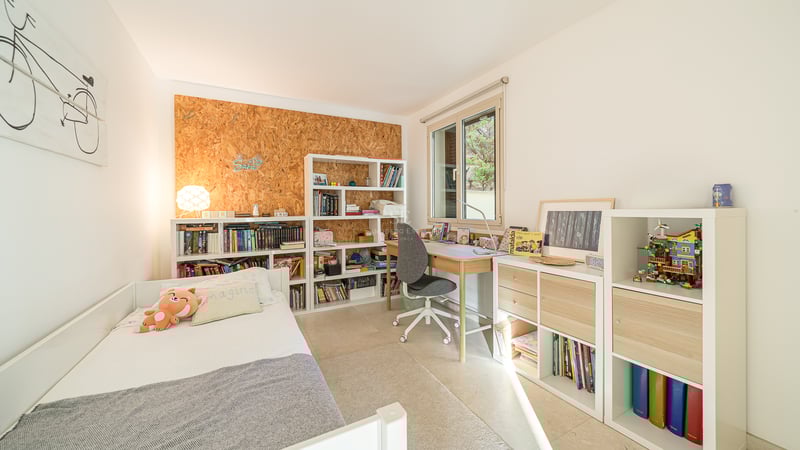 Barnrum med vit säng, skrivbord i trä och bokhyllor fyllda med böcker under ett ljust fönster.
