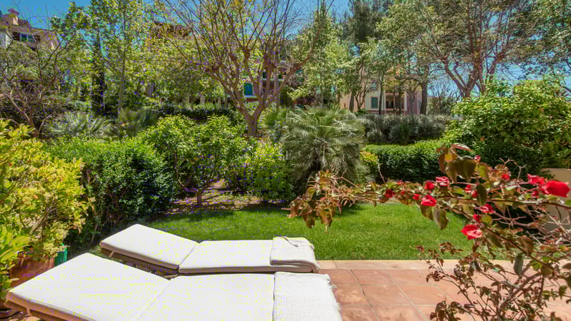 Two white sun loungers on a terracotta terrace overlooking a lush green garden with trees and flowering plants.