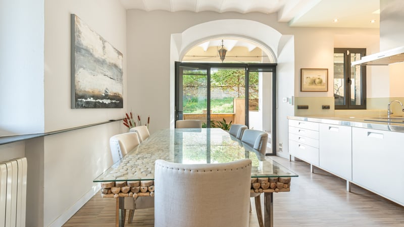 Ljus matsal med ett glasbord på en bas av trästockar, vita stolar och ett valv mot uteplatsen.