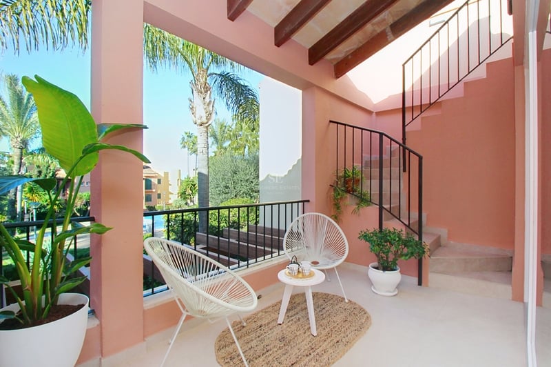 Laxrosa övertäckt terrass med vita Acapulco-stolar, träbjälkar och utsikt över poolområdet.