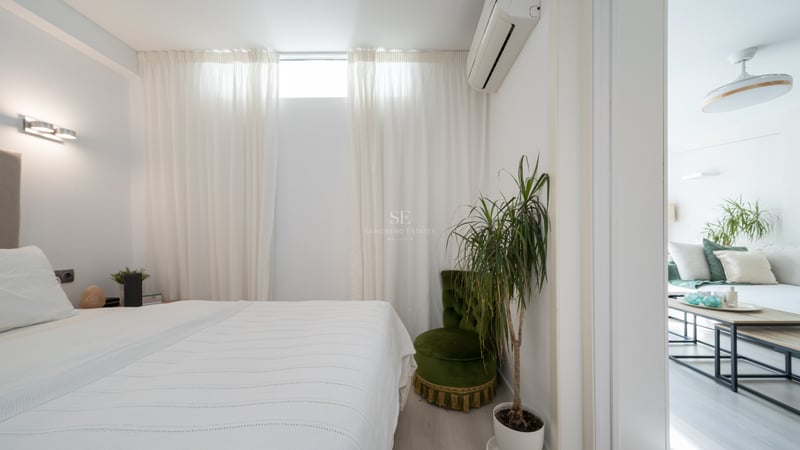 Clean white bedroom featuring a bed, green velvet chair, indoor plant, and a doorway looking into the living room.
