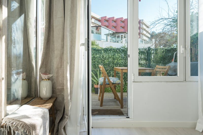 View through a glass door to a wooden terrace with outdoor furniture and a privacy hedge.