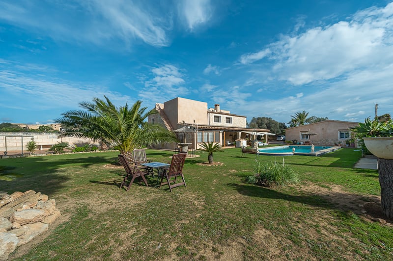 Extérieur d'une villa rustique avec pelouse, palmiers, piscine privée et mobilier de jardin sous un ciel bleu.