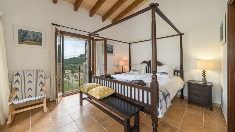 Chambre principale avec lit à baldaquin, poutres en bois, sol en terre cuite et balcon donnant sur les collines.