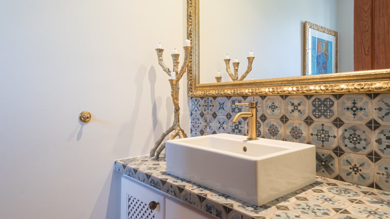 Close-up of a white vessel sink on a patterned tile countertop with a gold faucet and ornate gilded mirror.