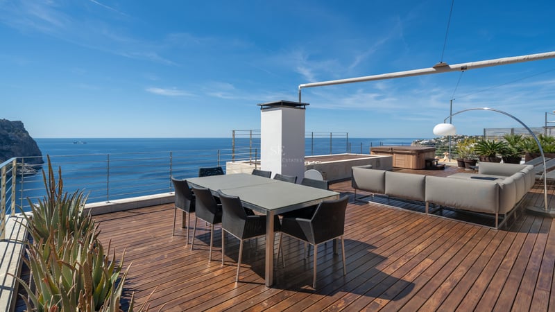 Modern rooftop terrace with wooden decking, dining table, lounge area, and glass railings overlooking the blue sea.