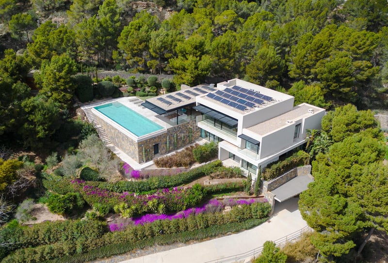Aerial view of a modern white villa with a rectangular infinity pool, solar panels, and surrounding pine forest.