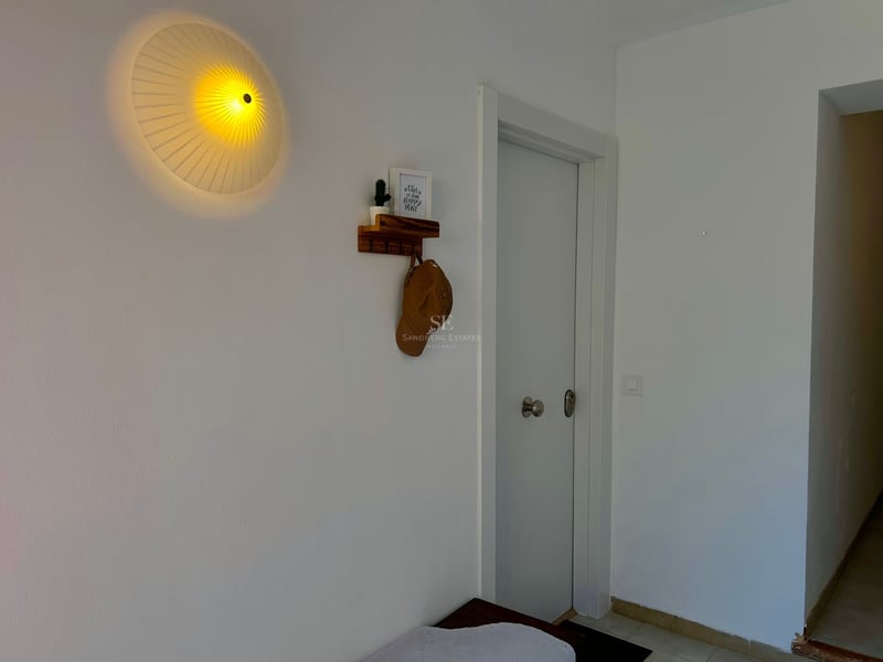 A white hallway featuring a round pleated wall lamp, a small wooden shelf with a hat, and a white door.