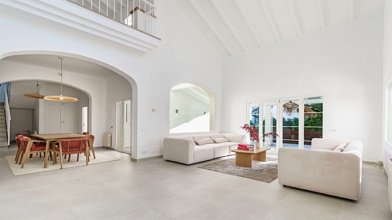 Rymligt vitt vardagsrum med högt i tak, bjälkar, beige soffor och matplats genom en rundbåge.