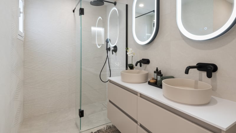 Modern bathroom with two beige vessel sinks, backlit oval mirrors, and a glass shower with textured wavy tiles.