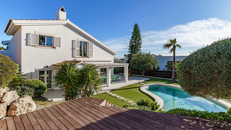 Vit tvåvåningsvilla med en njurformad pool, trädäck och välskött trädgård under klarblå himmel.