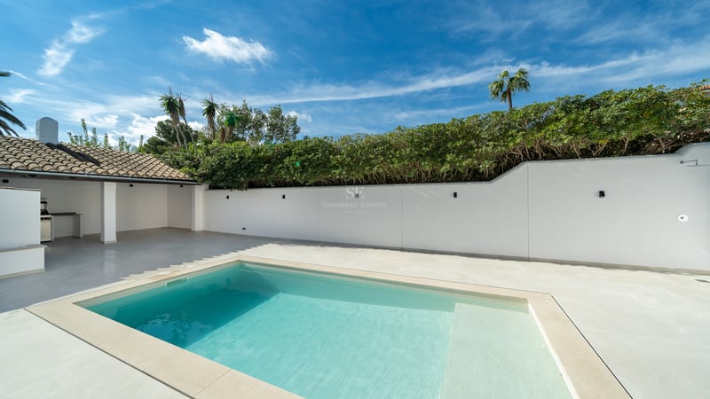 Piscine turquoise sur terrasse blanche avec mur d'intimité, haies vertes et cuisine d'été sous ciel bleu.