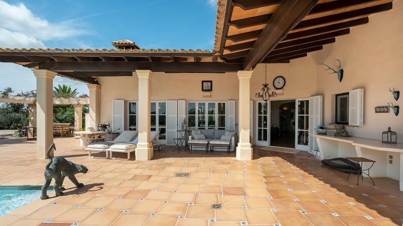 Terrasse en terre cuite avec poutres en bois sombre, piliers blancs, chaises longues et volets blancs sous le soleil.