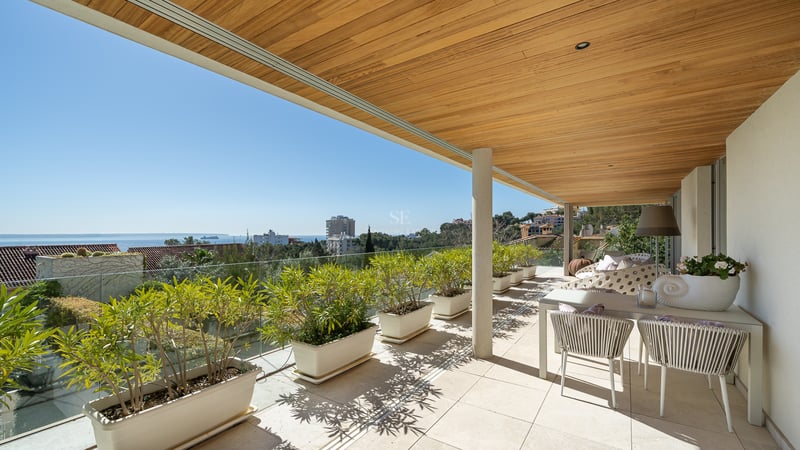 Überdachte Terrasse mit Holzdecke, Glasgeländer und weißen Pflanzgefäßen mit Blick auf die Stadt.