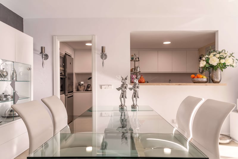 Zeitgenössisches Esszimmer mit Glastisch, weißen Stühlen und Blick in eine moderne weiße Küche durch eine Durchreiche.