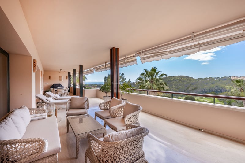Grand balcon couvert avec mobilier en osier offrant une vue sur une vallée verdoyante et la mer par temps clair.