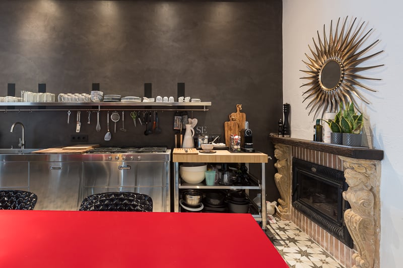 Cuisine moderne avec meubles en acier inoxydable, mur sombre texturé, table rouge et cheminée en pierre sculptée.