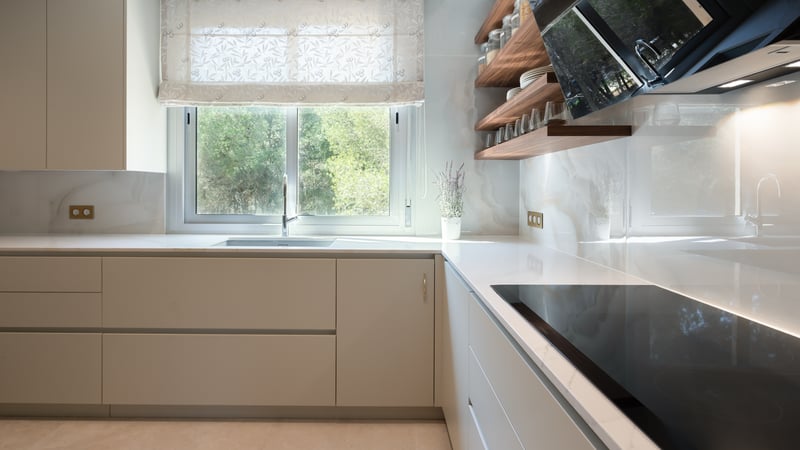 Minimalist off-white kitchen with marble-effect backsplash, wooden shelves, and a window overlooking lush greenery.