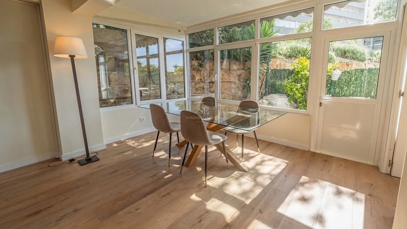 Comedor luminoso con mesa de cristal, sillas tapizadas en beige y amplios ventanales que dan a un jardín mediterráneo.