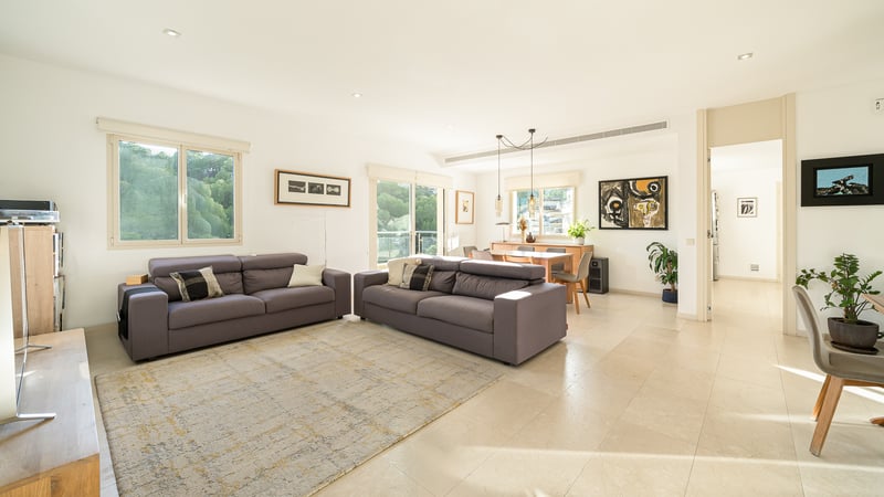 Rymligt och ljust vardagsrum med två grå soffor, ljusa klinkergolv och matplats i bakgrunden med stora fönster.