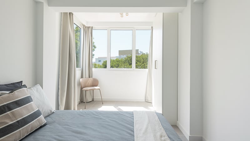 Une chambre lumineuse et moderne avec un lit gris, des placards intégrés blancs et une fenêtre donnant sur des arbres.