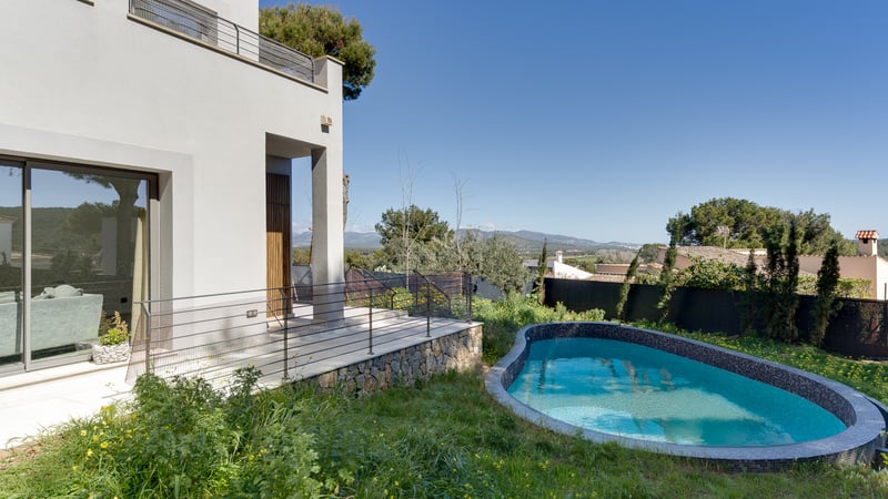 Modern vit villa med en kurvad blå mosaikpool, stenvägg och bergsutsikt under en klarblå himmel.