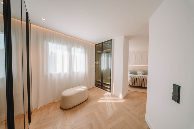 Dressing lumineux avec parquet en chêne, pouf beige et armoire vitrée menant à une chambre.