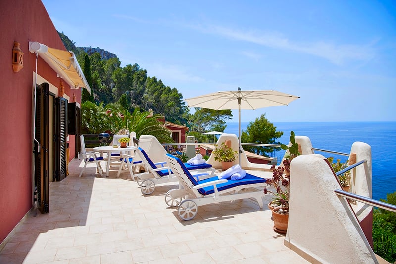 Sunny terrace with blue loungers, white umbrella, and panoramic views of the Mediterranean Sea and cliffs.