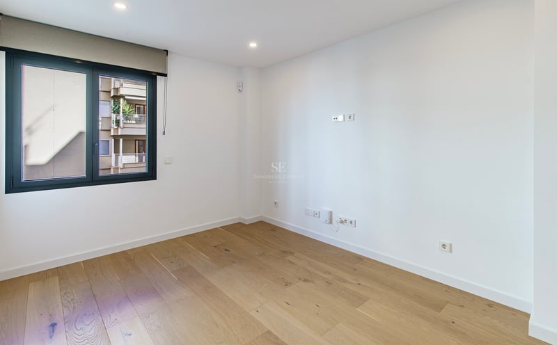 Pièce vide et lumineuse avec parquet en bois clair, murs blancs et une grande fenêtre avec store.