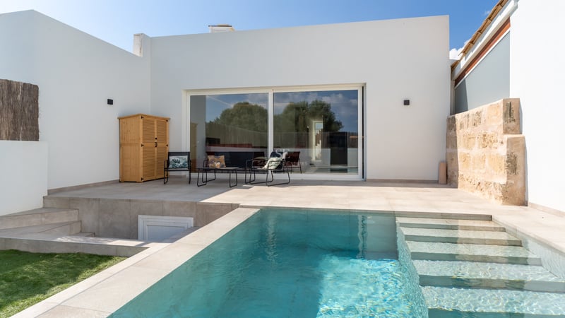 Rectangular turquoise swimming pool with stone steps integrated into a light stone terrace next to a white villa facade.