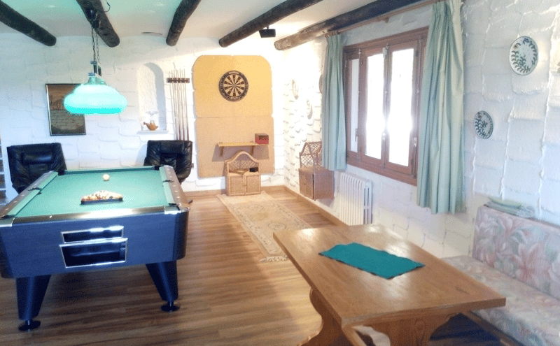 Sala de ocio con mesa de billar, diana, paredes de piedra blanca y vigas de madera oscura.
