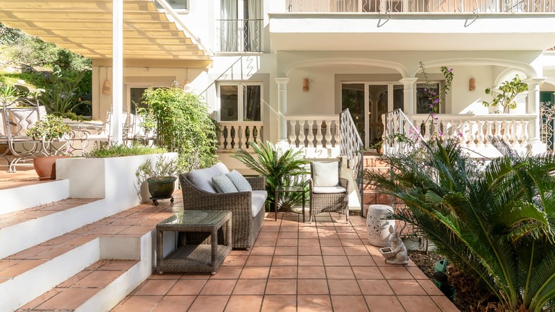 Sun-drenched terracotta tile terrace with wicker lounge furniture and lush greenery against a classic white villa facade.