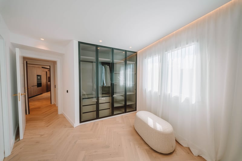 Dressing moderne avec parquet en chevrons, armoire vitrée et pouf central près d'une fenêtre.