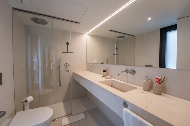 Modernt badrum med handfat i beige sten, duschvägg i glas, stor spegel och linjär belysning.