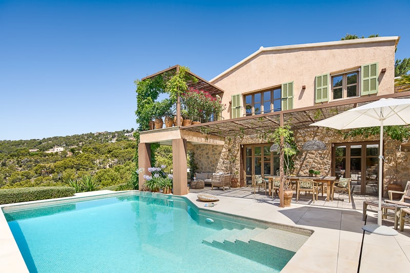 Exterior view of a stone villa featuring a turquoise pool, outdoor dining area, and panoramic hillside views.