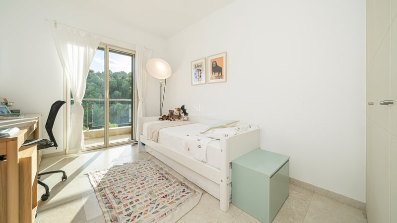 Vitt sovrum med enkelsäng, skrivbord, balkongdörr och mönstrad matta.