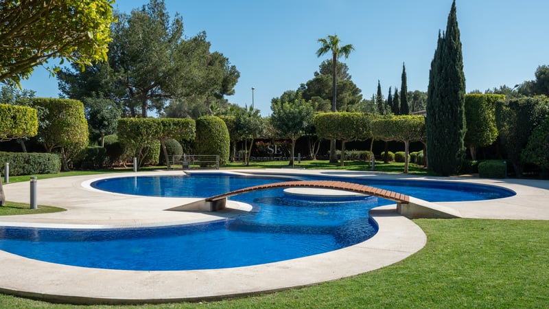 An expansive multi-lobed blue swimming pool with a wooden footbridge, surrounded by manicured lawns and mature trees.