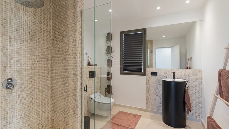 Modernt badrum med mosaikkakel, duschvägg i glas, mattsvart pelarhandfat och vägghängd toalett.