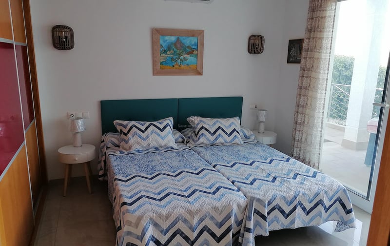 Chambre avec lits jumeaux, tête de lit bleu canard et grande baie vitrée ouvrant sur une terrasse ensoleillée.