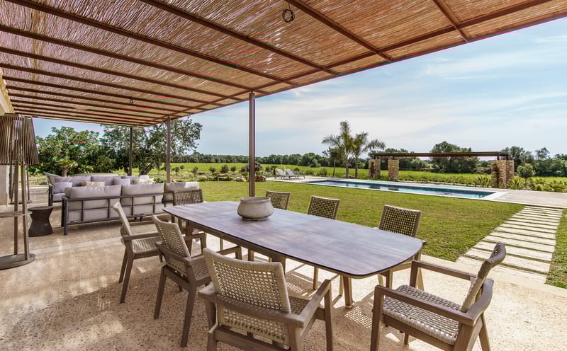 Großer Holzesstisch unter einer Schilfpergola auf einer Steinterrasse mit Blick auf Pool und Rasen.