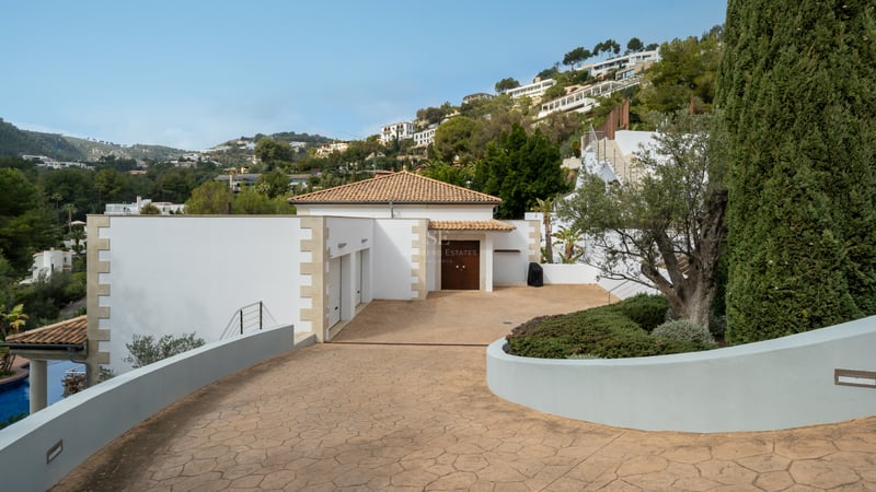 Une allée privée pavée menant à une villa blanche avec toit en terre cuite, double garage et végétation luxuriante.