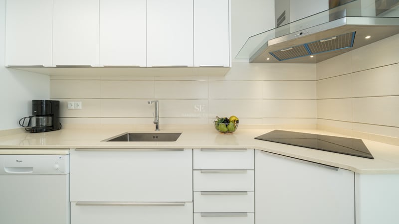 Close-up of a modern white kitchen featuring a stainless steel sink, glass hood, and ceramic cooktop.