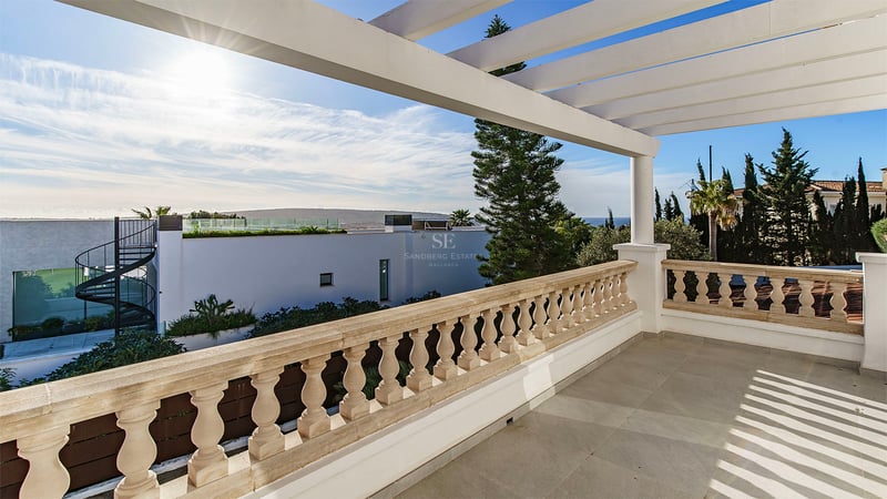 Rymlig terrass med stenbalustrad och vit pergola med utsikt över kustlandskapet och en klarblå himmel.