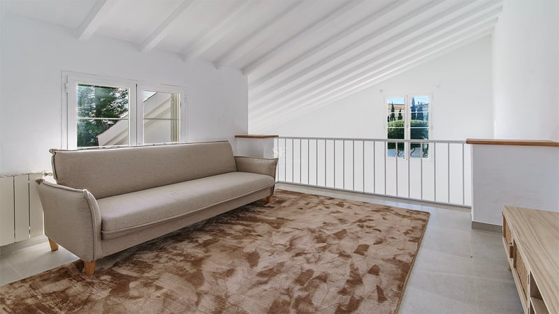 Vardagsrum på loftet med beige soffa, mönstrad brun matta och vitmålade takbjälkar i trä.